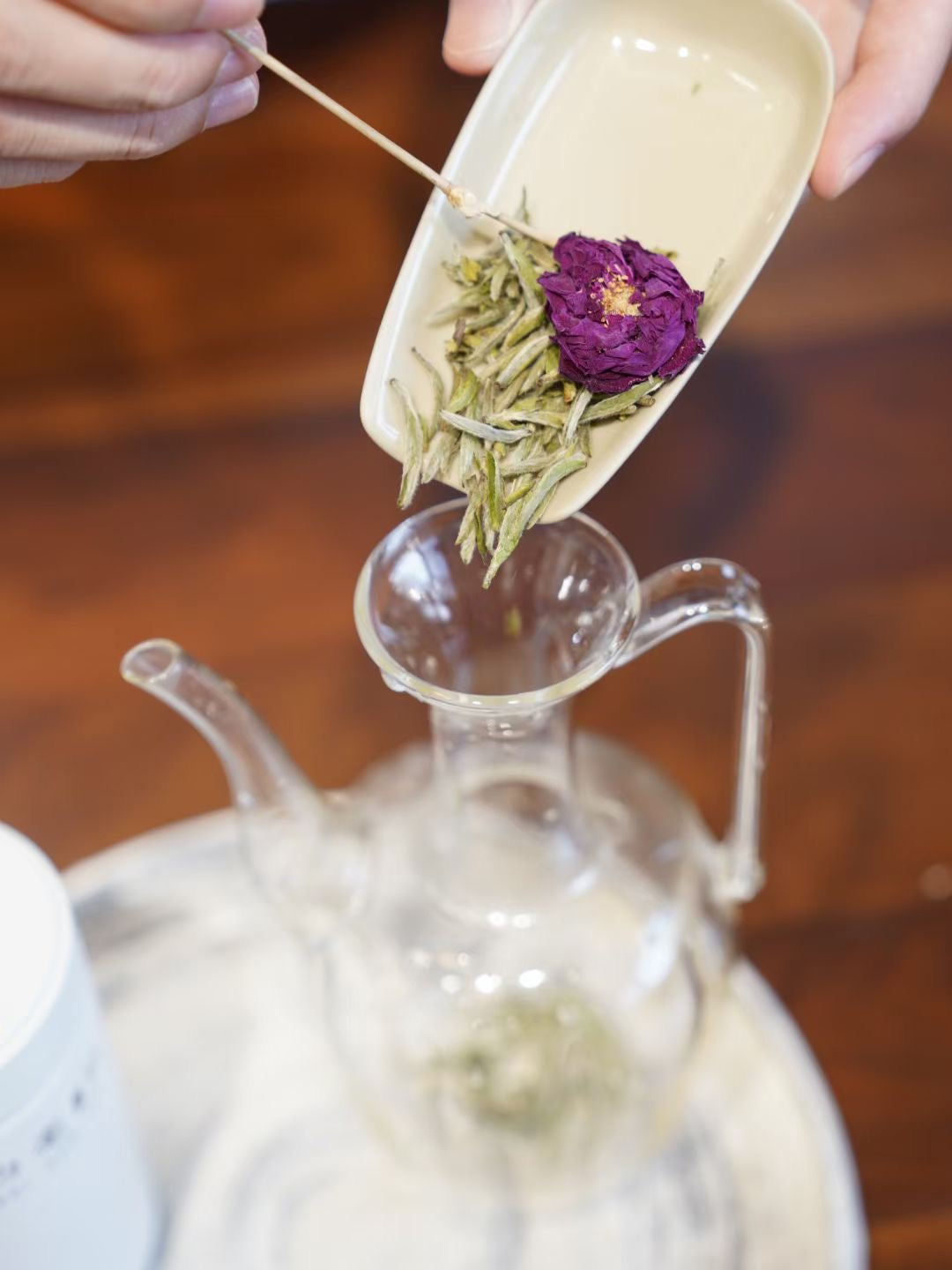 Tea leaves and a purple flower being added to a glass teapot.