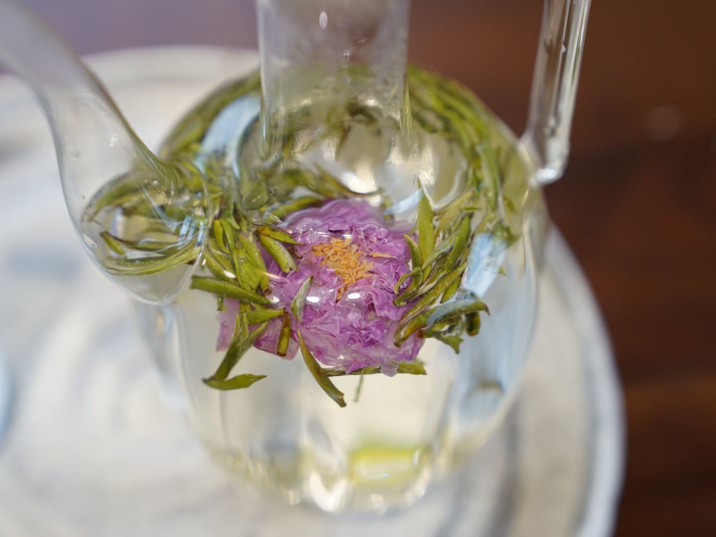 Clear glass teapot with pink flower and green leaves inside on a white surface.