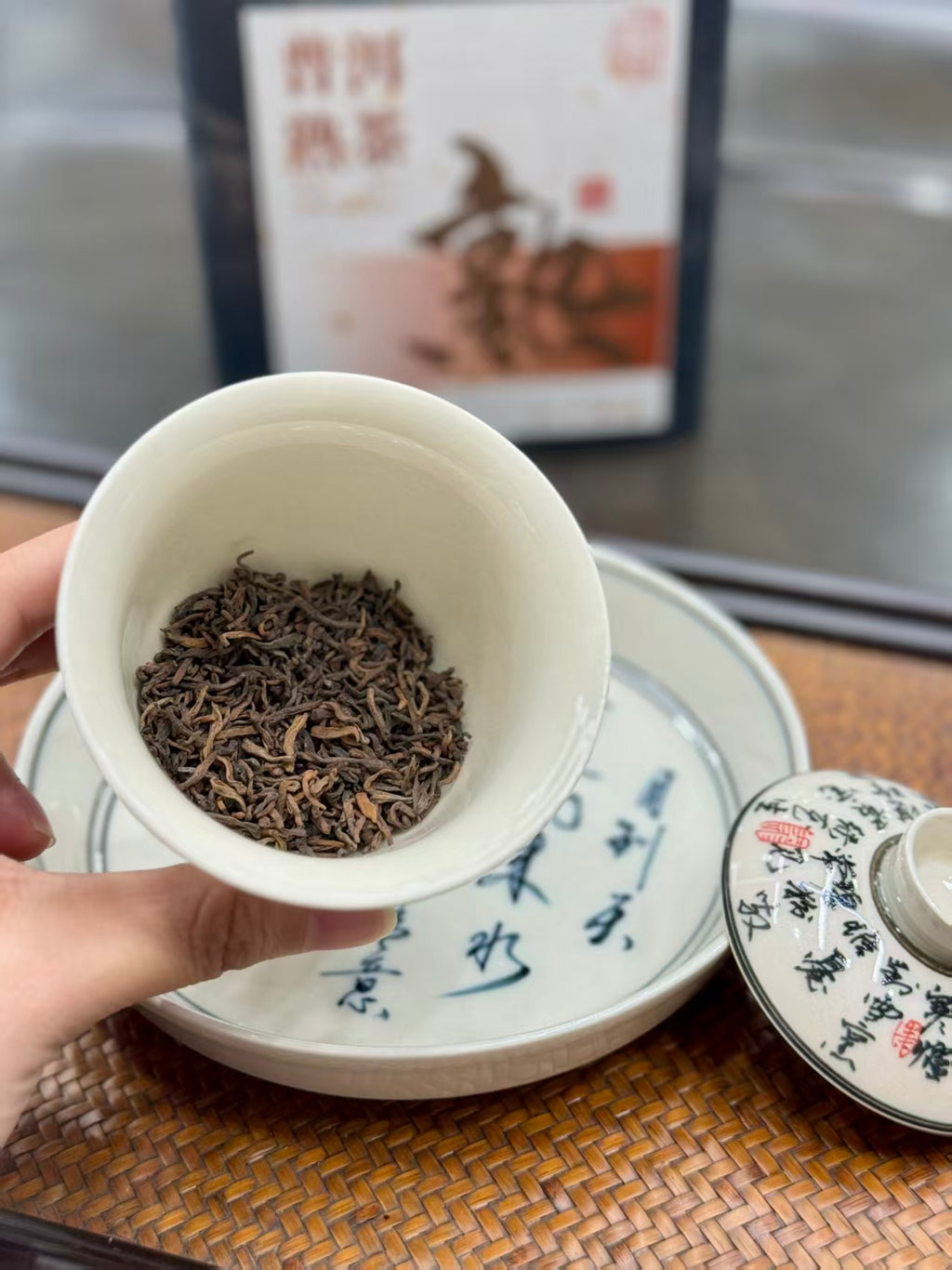 A hand holds a small white ceramic teacup filled with loose-leaf tea leaves, with a matching teacup lid and saucer in the foreground and a tea package blurred in the background. Ten years in the making. Ready in a minute.
Aged ripe pu-erh, loose and ready to brew. Two tins, one to open now, one to keep deepening.
