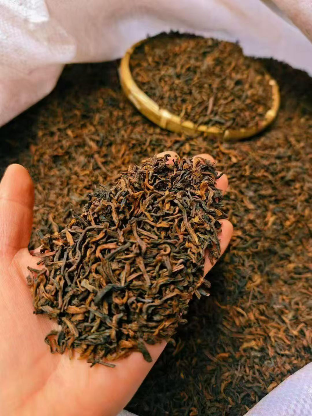 A close-up of a hand holding a small pile of dark, curly tea leaves, with more tea visible in a woven tray and spread out behind it inside a white sack. Ten years in the making. Ready in a minute.
Aged ripe pu-erh, loose and ready to brew. Two tins, one to open now, one to keep deepening.