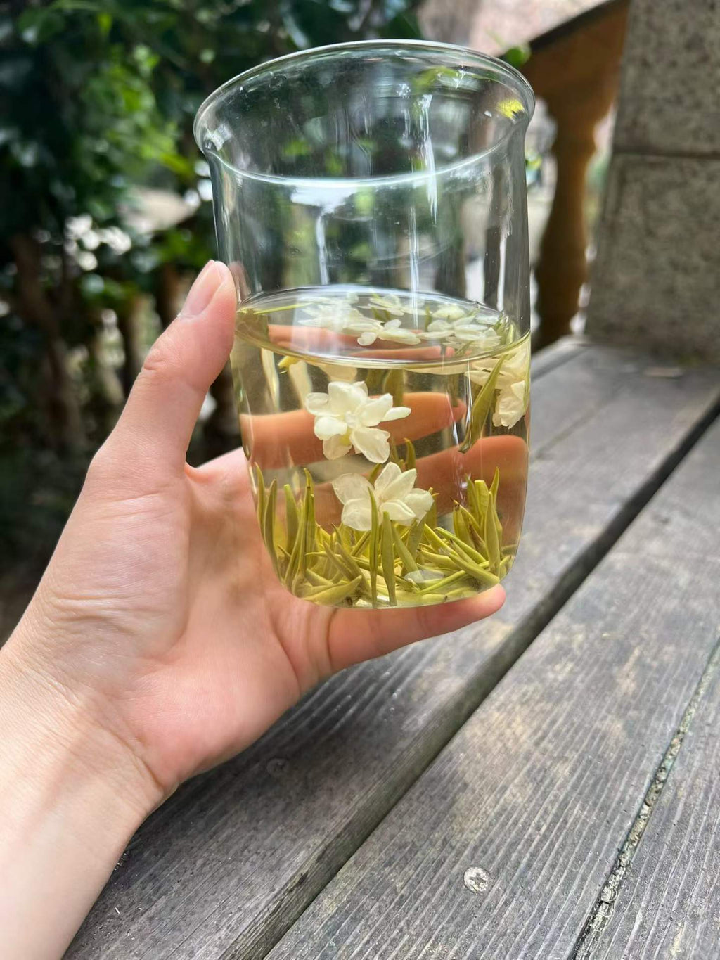 Hand holding a glass of clear liquid with floating flowers on a wooden . Exhale — 2026 fresh harvest jasmine needle tea for anxiety and stress relief. Premium spring buds, intensely fragrant, cooling golden liquor. Breathe out.