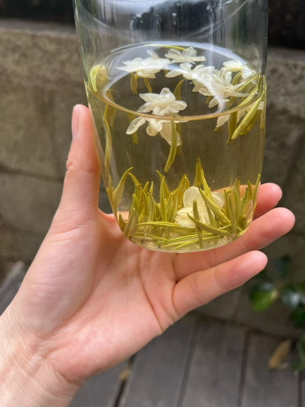 Hand holding a glass of green tea with flowers against a blurred background. Exhale — 2026 fresh harvest jasmine needle tea for anxiety and stress relief. Premium spring buds, intensely fragrant, cooling golden liquor. Breathe out.