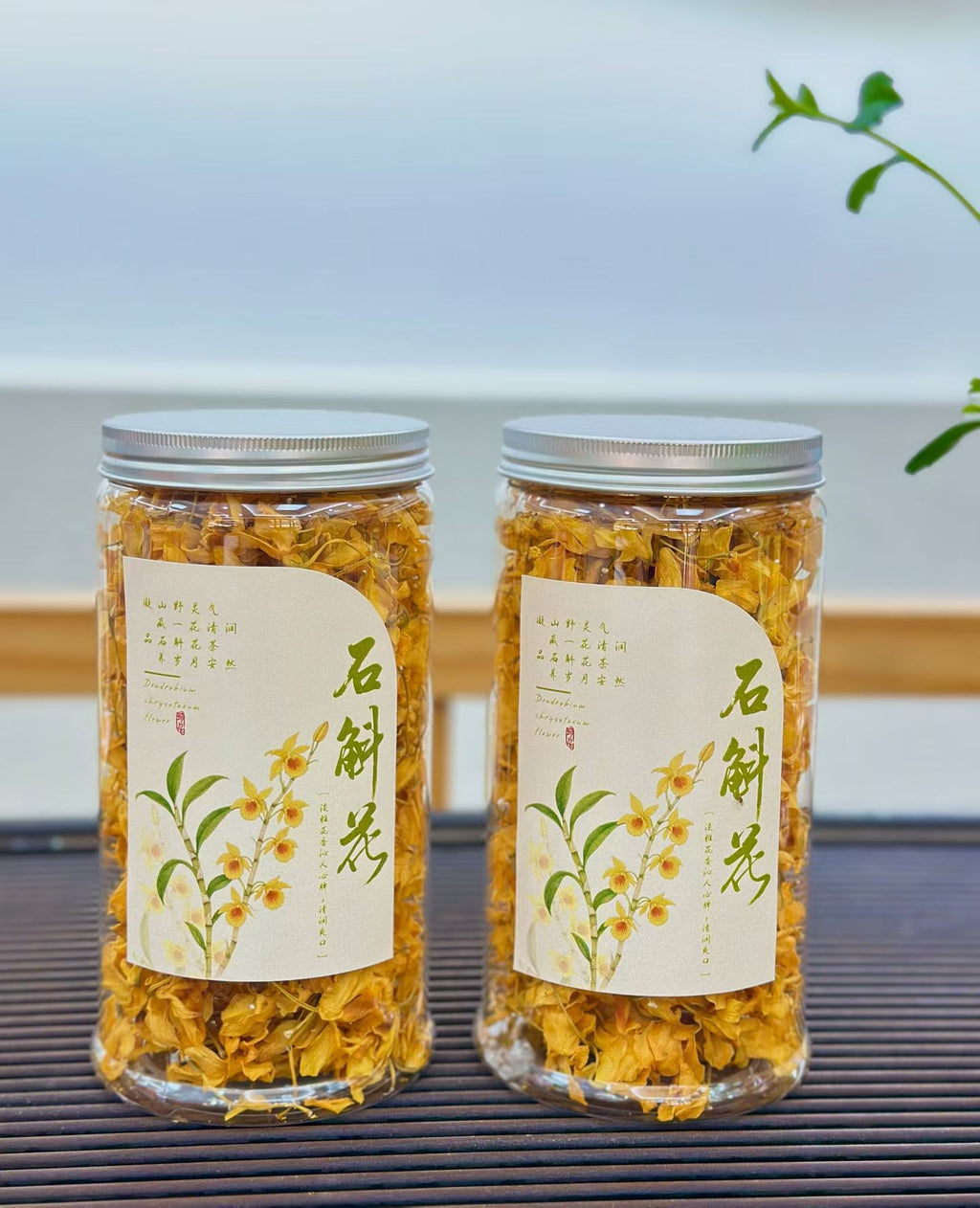 Two clear glass jars filled with dried yellow flowers, sealed with silver lids and labeled with cream paper stickers featuring green calligraphy and floral illustrations, set on a dark slatted surface with a soft blurred background.