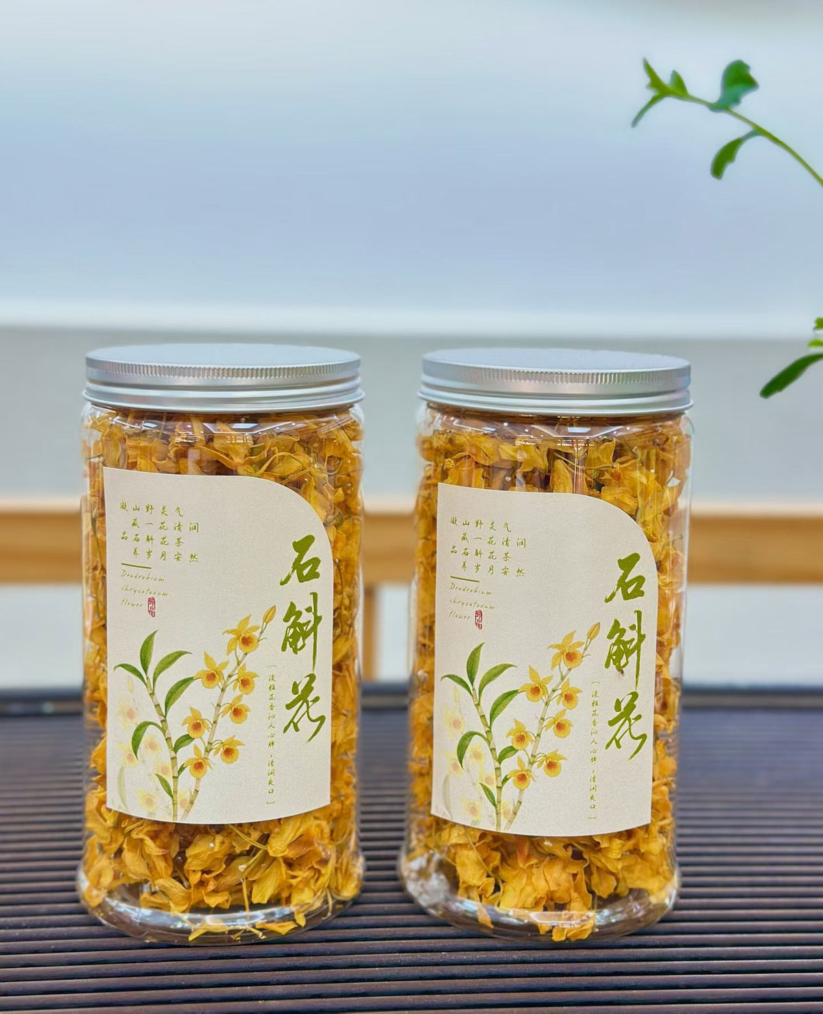 Two clear glass jars filled with dried yellow flowers, sealed with silver lids and labeled with cream paper stickers featuring green calligraphy and floral illustrations, set on a dark slatted surface with a soft blurred background.