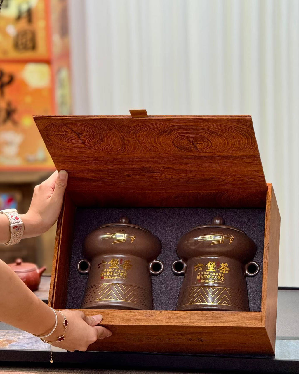 A wooden gift box with two brown ceramic tea jars inside, held open by a person's hands in a warm indoor setting.