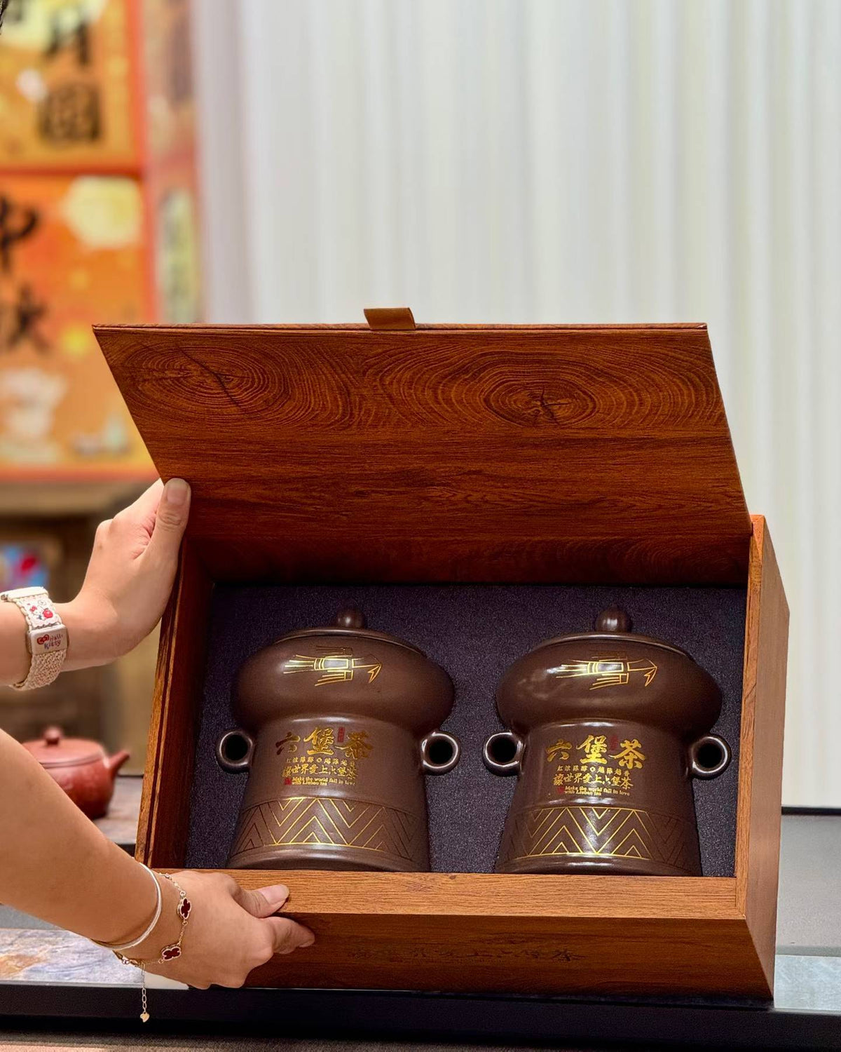 A wooden gift box with two brown ceramic tea jars inside, held open by a person's hands in a warm indoor setting.