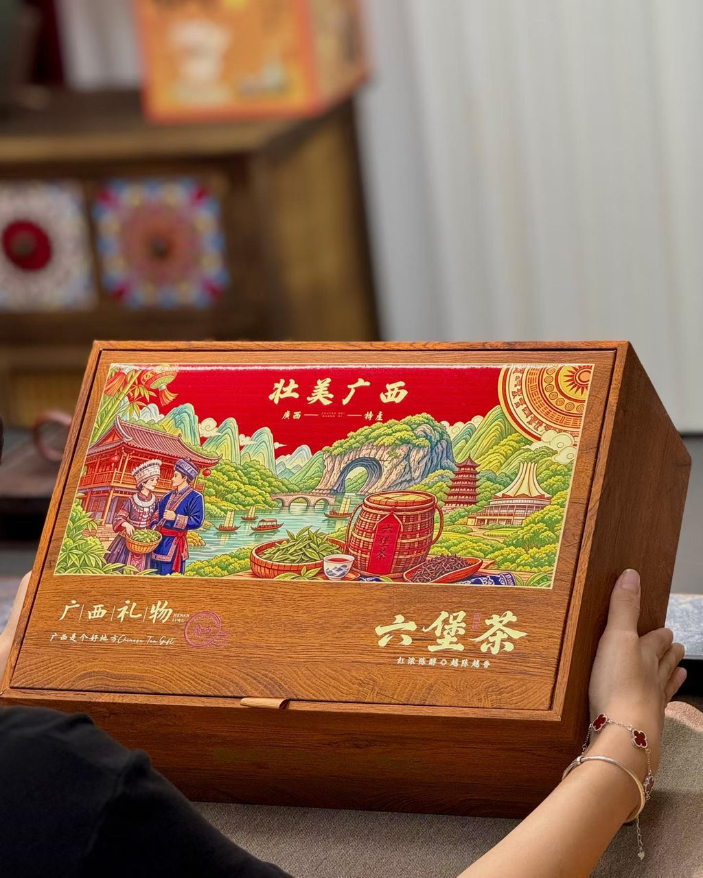 A wooden gift box for Guangxi six-bottle tea, held by a person's hands in a home setting. The lid features a colorful, detailed illustration of a traditional scene with people, mountains, and tea barrels, with Chinese text.