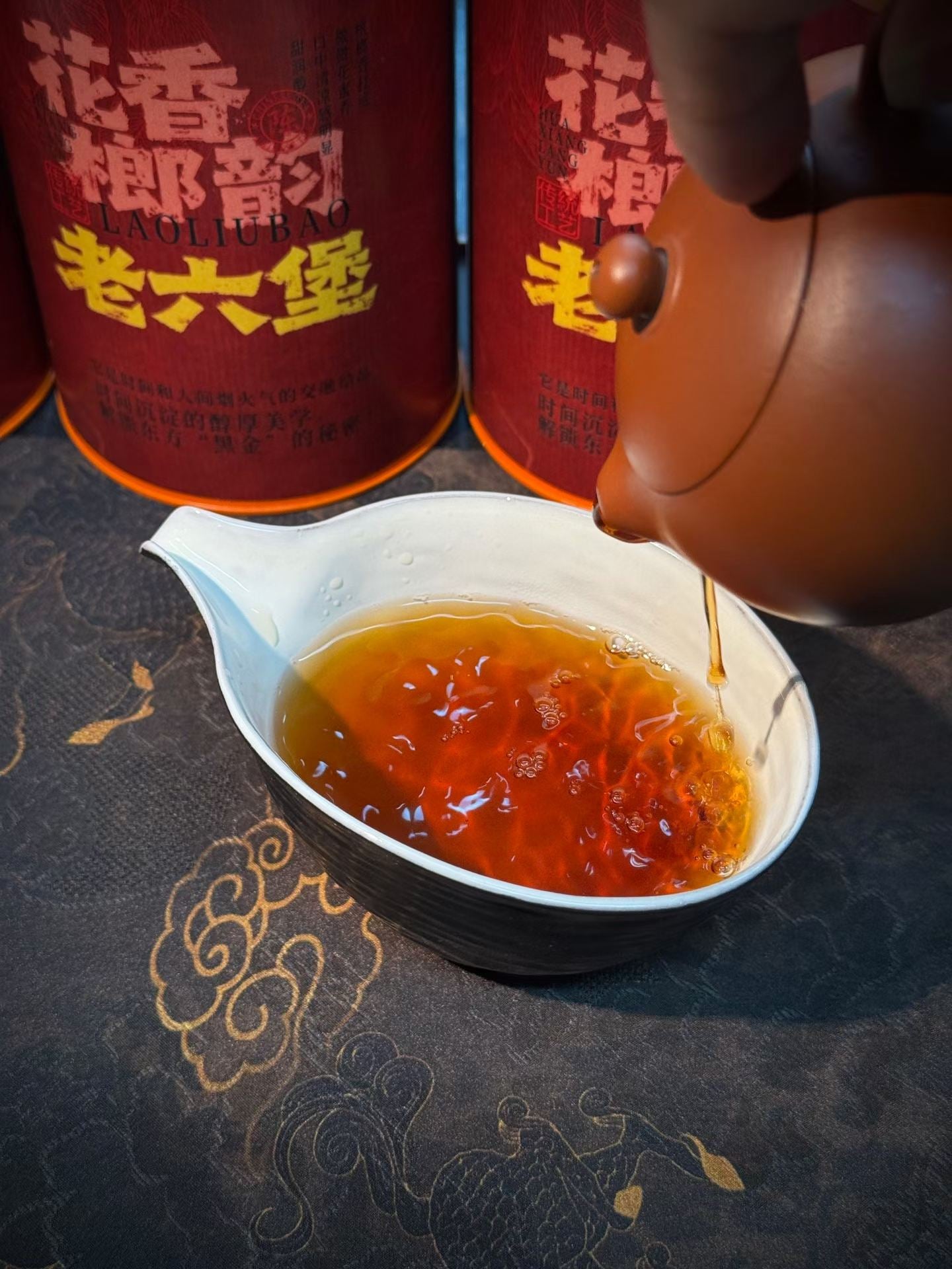 Tea being poured from a teapot into a white cup with red tea canisters in the background. Liu Bao aged dark tea from Wuzhou, Guangxi. National intangible heritage workshop, bamboo basket storage. Supports digestion and gut balance. For drinkers and collectors.