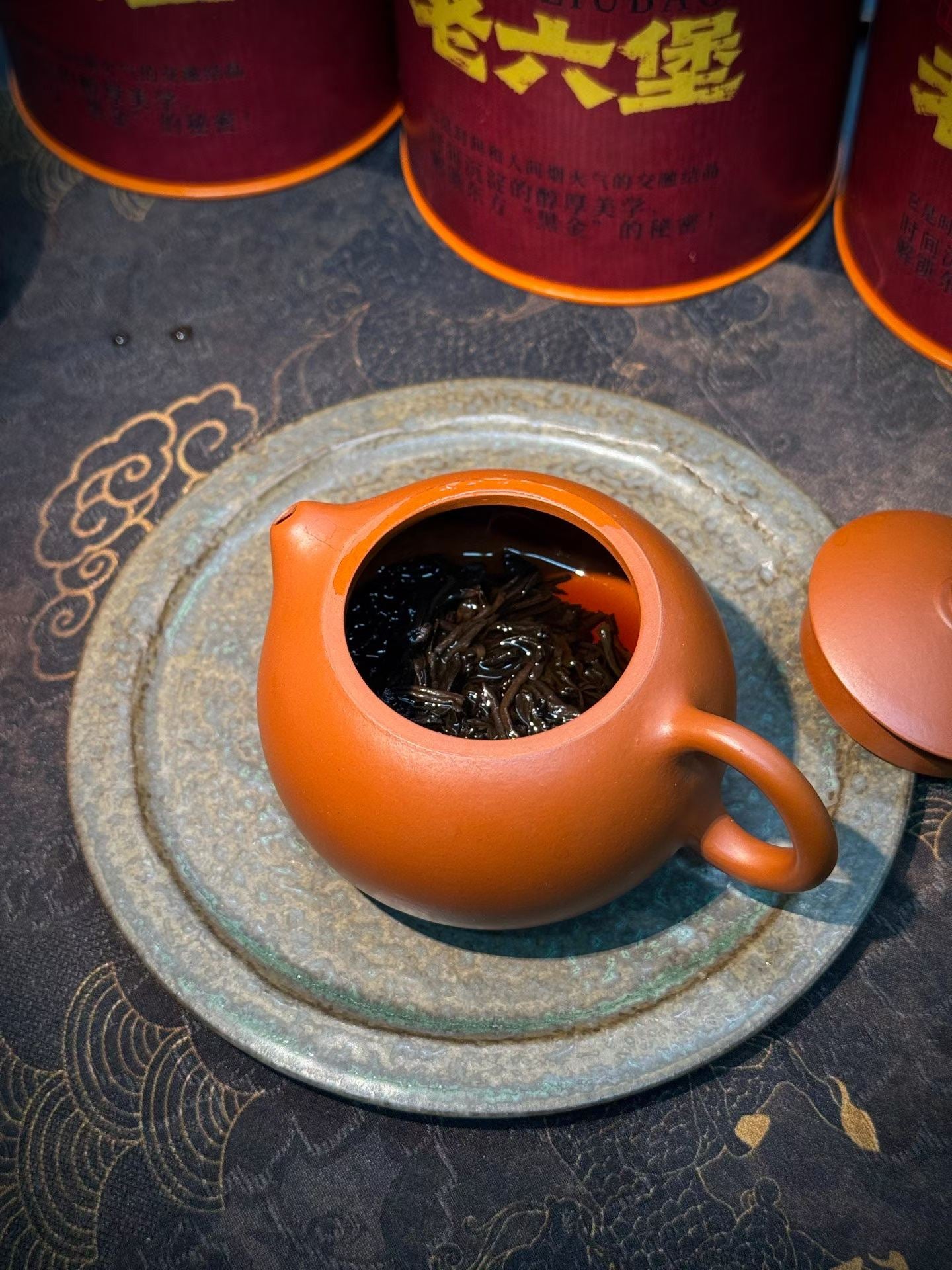 Orange ceramic teapot with tea leaves on a textured surface with red containers in the background.Liu Bao aged dark tea from Wuzhou, Guangxi. National intangible heritage workshop, bamboo basket storage. Supports digestion and gut balance. For drinkers and collectors.
