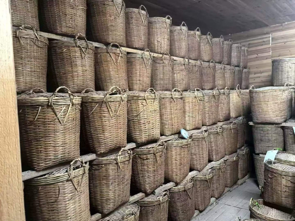Row of wicker baskets stacked against a wall in a storage room. Liu Bao aged dark tea from Wuzhou, Guangxi. National intangible heritage workshop, bamboo basket storage. Supports digestion and gut balance. For drinkers and collectors.
