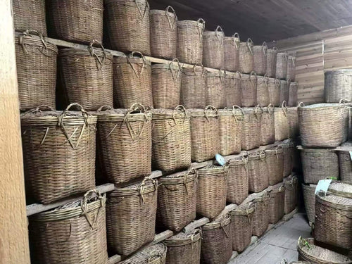 Row of wicker baskets stacked against a wall in a storage room. Liu Bao aged dark tea from Wuzhou, Guangxi. National intangible heritage workshop, bamboo basket storage. Supports digestion and gut balance. For drinkers and collectors.