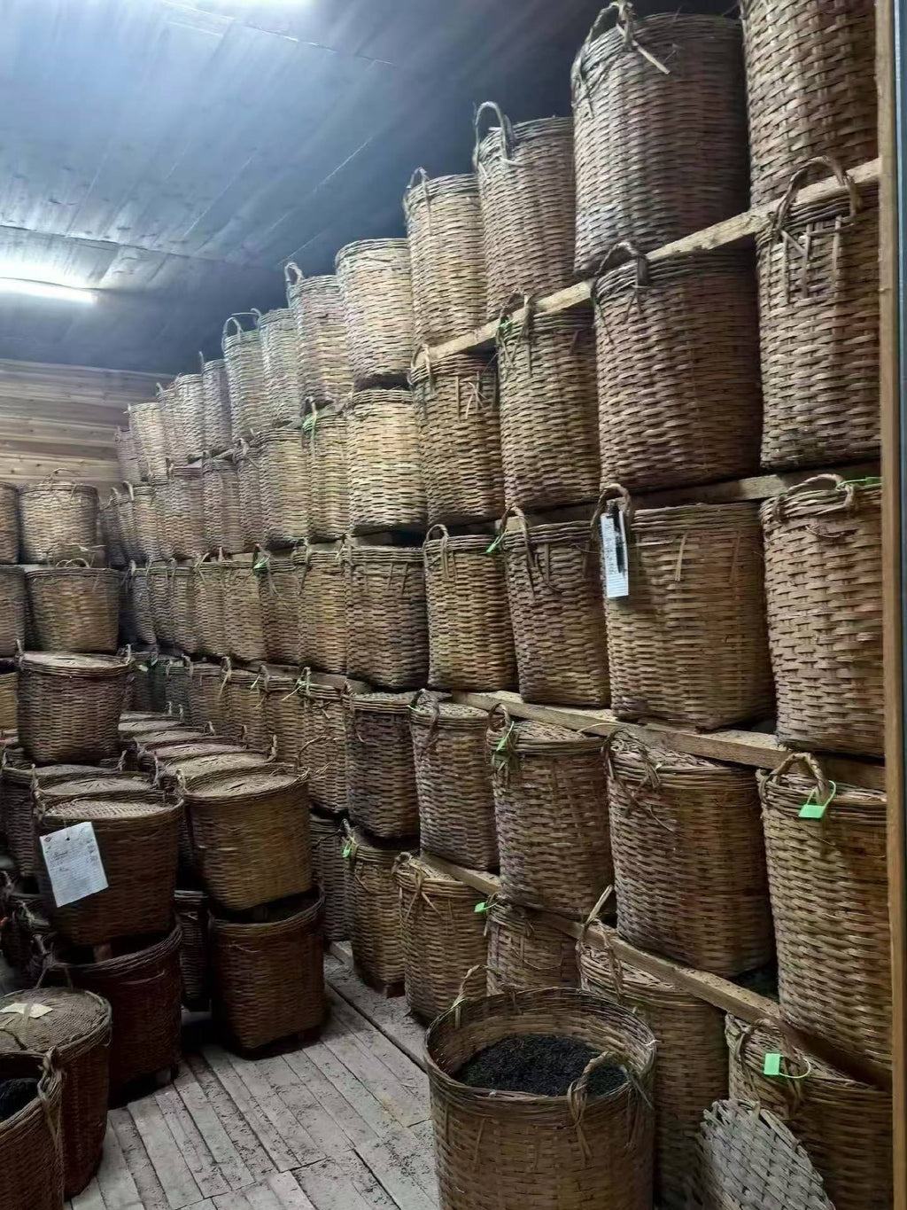 Stacks of wicker baskets in a storage area with wooden floors and a concrete wall. Liu Bao aged dark tea from Wuzhou, Guangxi. National intangible heritage workshop, bamboo basket storage. Supports digestion and gut balance. For drinkers and collectors.
