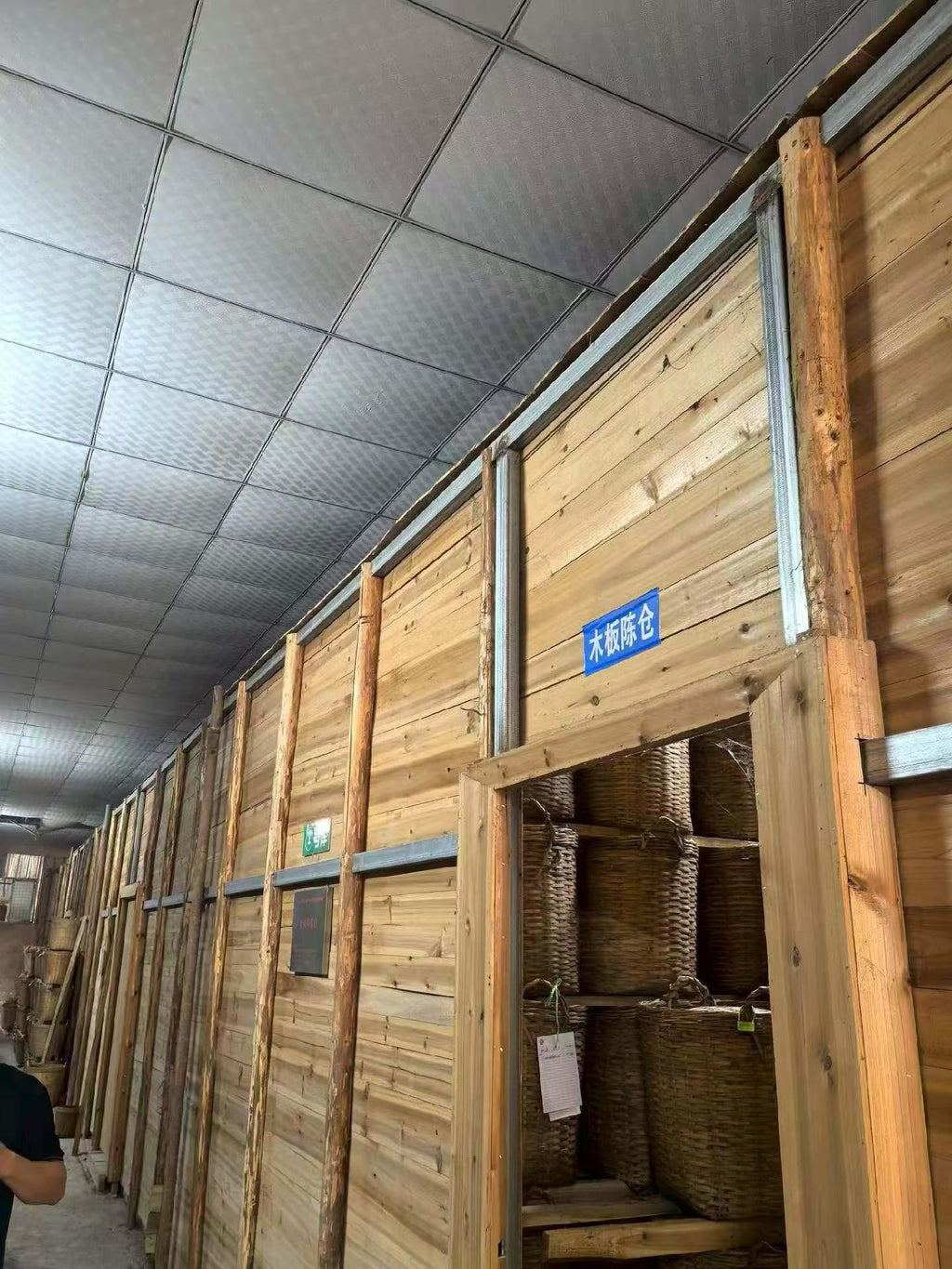 Row of wooden crates stacked against a wall in an indoor setting. Liu Bao aged dark tea from Wuzhou, Guangxi. National intangible heritage workshop, bamboo basket storage. Supports digestion and gut balance. For drinkers and collectors.