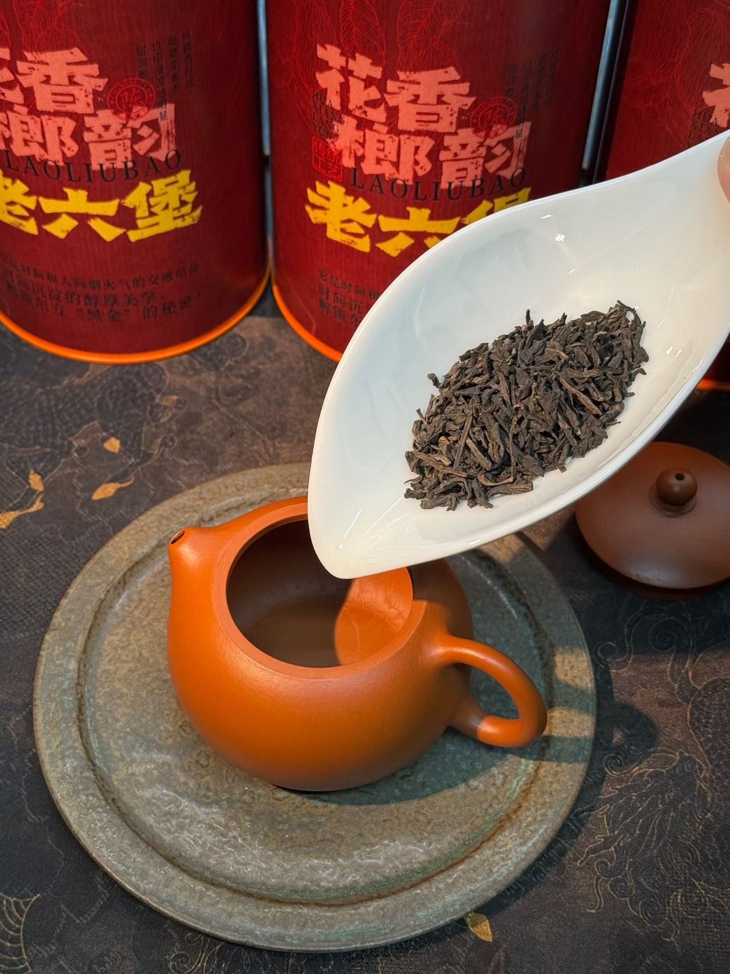 Tea leaves being scooped into a teacup with tea tins in the background. Liu Bao aged dark tea from Wuzhou, Guangxi. National intangible heritage workshop, bamboo basket storage. Supports digestion and gut balance. For drinkers and collectors.