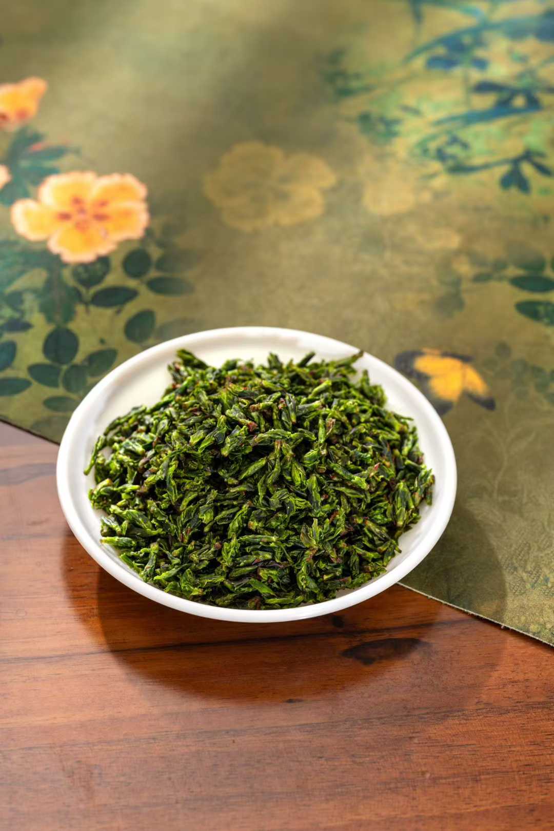 Small-Leaf Kuding · 2026 First Spring | Pure Rice-Bud Harvest, White bowl filled with green tea leaves on a wooden surface with a floral-patterned cloth in the background.