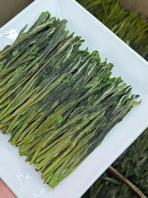 Dried green tea leaves on a white plat. Taiping Houkui hand-pressed green tea from Huangshan's high mountains. First-flush leaves, intangible-heritage craft, sword-straight blades, clear emerald liquor. Orchid-soft and sweet.