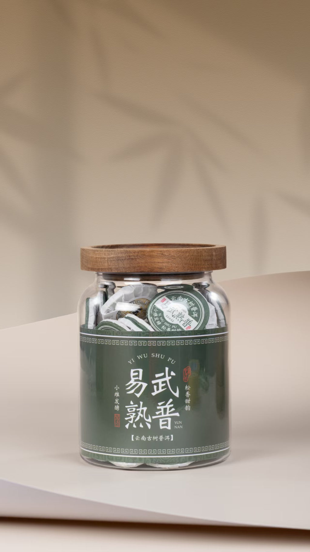 A clear glass jar filled with round tea discs, topped with a wooden lid and wrapped in a dark green label with Chinese characters, set on a beige surface against a beige background with soft leaf shadows. The leaves for this tea come from Yiwu's old trees. They are fermented in small piles, slowly and carefully, so the tea stays clean and gentle. Then each portion is pressed into a small round cake, about the size of a coin.