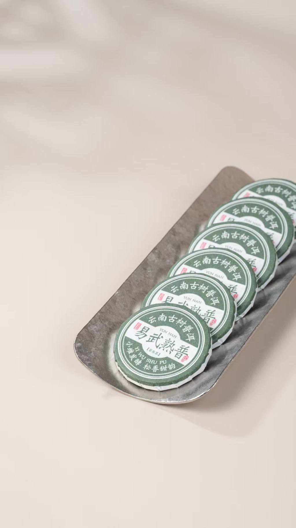 A row of round green tea cakes with white labels sits on a silver metal tray against a soft beige background. The leaves for this tea come from Yiwu's old trees. They are fermented in small piles, slowly and carefully, so the tea stays clean and gentle. Then each portion is pressed into a small round cake, about the size of a coin.