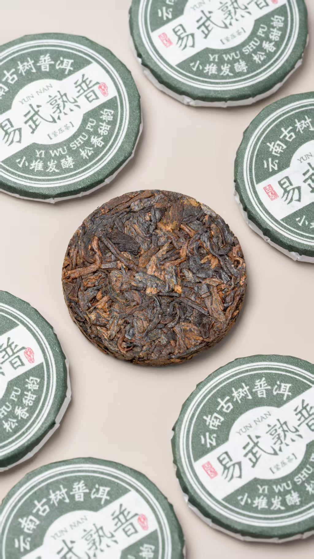 A single round dark brown compressed tea cake is centered among several green-and-white tea tin cans with Chinese text, arranged on a light beige background. The leaves for this tea come from Yiwu's old trees. They are fermented in small piles, slowly and carefully, so the tea stays clean and gentle. Then each portion is pressed into a small round cake, about the size of a coin.