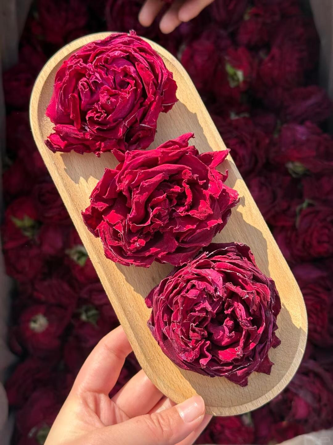 Three dried red flowers on a wooden tray held by a hand with a blurred background of more flowers. The deepest red Yunnan grows. In your cup.
