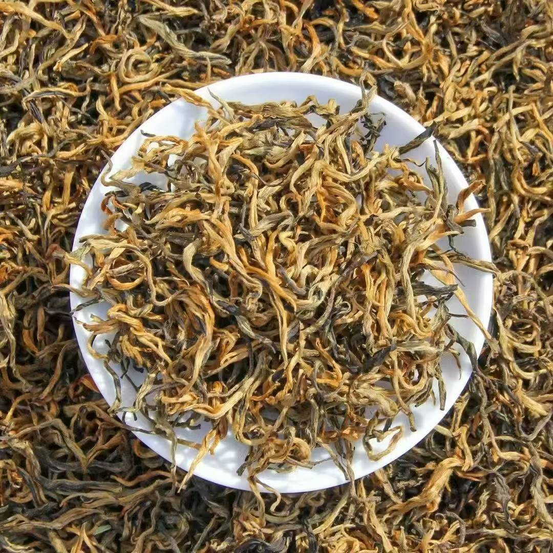 Dry tea leaves in a white bowl surrounded by more tea leaves on a textured surface. 2026 Spring Glutinous Aroma Golden Bud Black Tea – High Bud Content (70–80%)