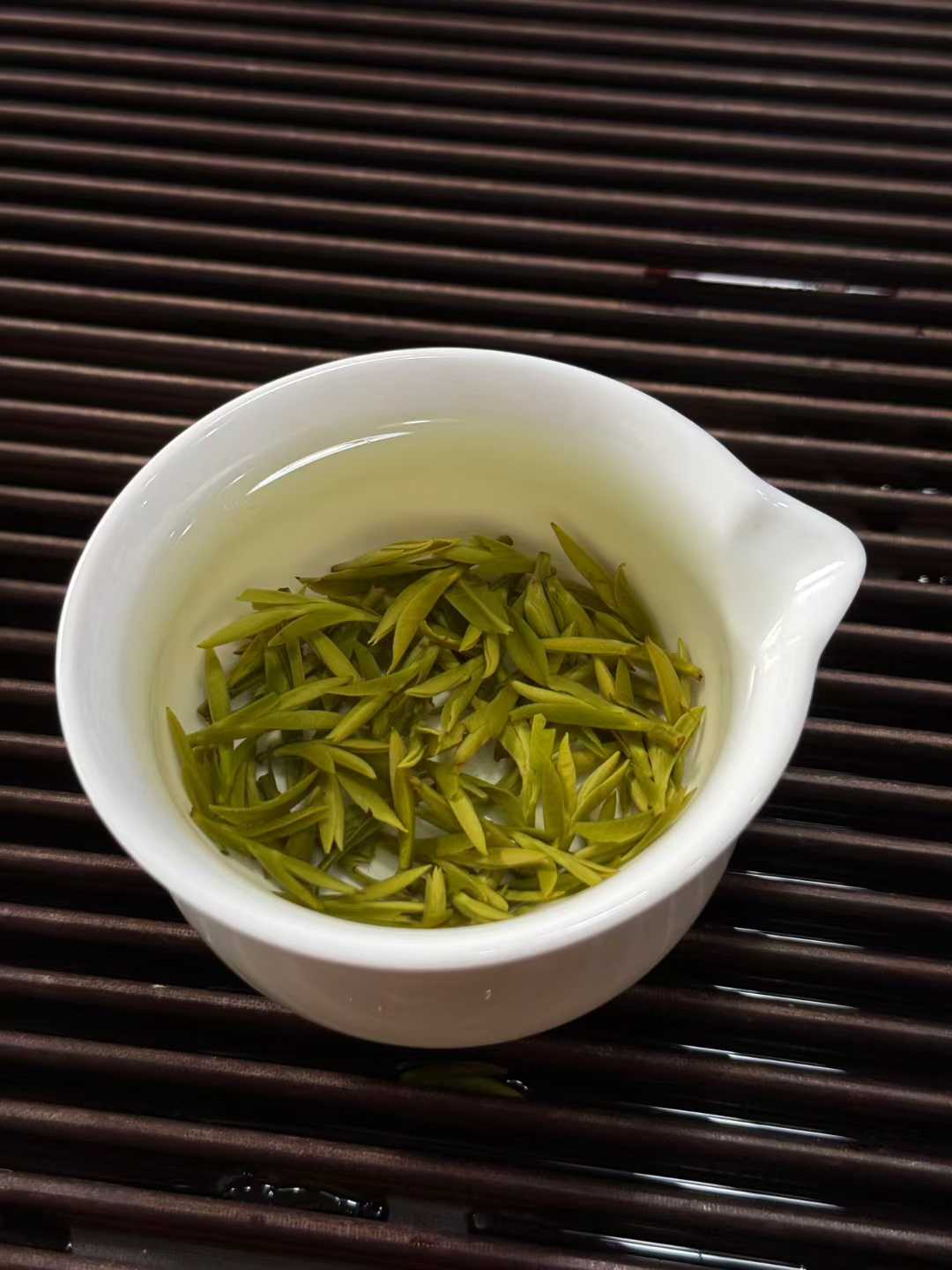 White cup with green tea leaves on a dark wooden surface. 2026 First Harvest Huangshan Maofeng – Early Spring Imperial Green Tea
