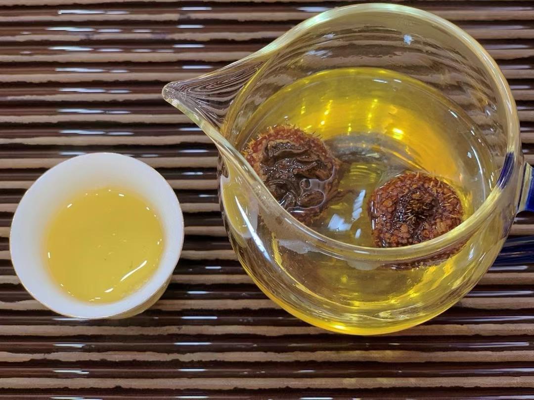 Glass teapot with tea leaves and a small white dish of tea on a woven surface. Ci li black tea — aged tea leaves brewed inside whole wild rose hip fruit from Guizhou. Natural vitamin C, golden liquor, creamy finish. Nothing added.