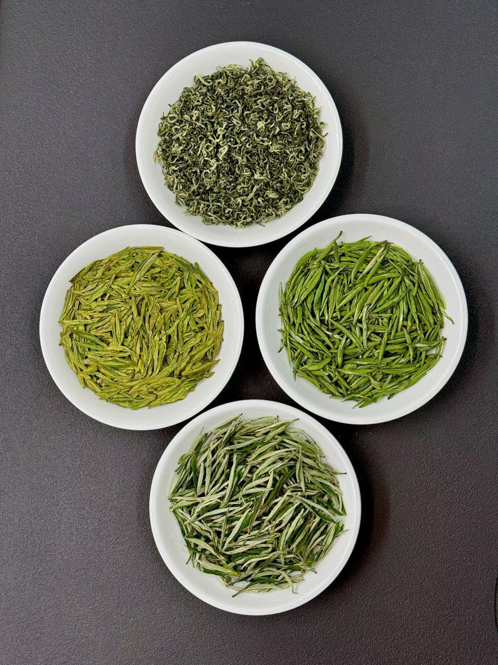 Four white bowls filled with different types of green tea leaves on a dark gray background. Four of China’s most iconic spring green teas — harvested during the precious pre-Qingming season, when tea buds are at their most tender, fragrant, and vibrant.