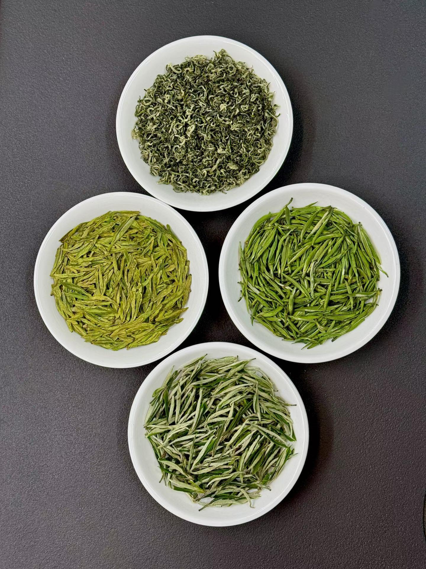 Four white bowls filled with different types of green tea leaves on a dark gray background. Four of China’s most iconic spring green teas — harvested during the precious pre-Qingming season, when tea buds are at their most tender, fragrant, and vibrant.