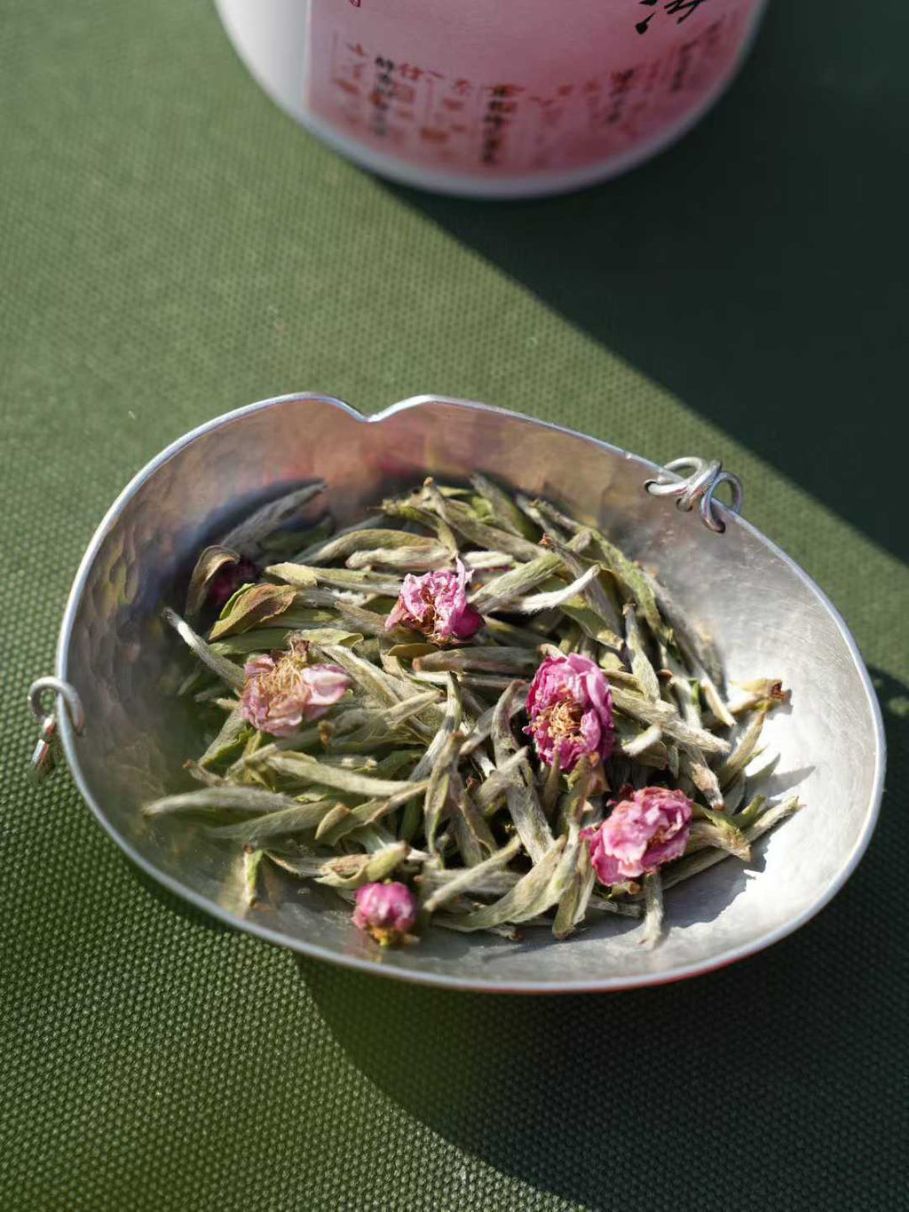 Silver teabag with green tea leaves and pink flowers on a green surface