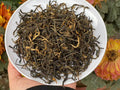 Dry tea leaves on a white plate held by a hand with a floral background. Yunnan nuoxiang black tea with a natural glutinous rice aroma. Mellow, warming, deeply comforting. A daily ritual tea for stress relief and quiet evenings.