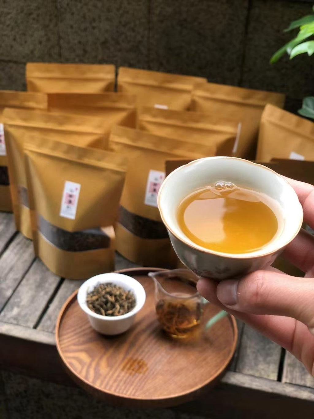 Person holding a teacup with tea, surrounded by tea packages and a small bowl of tea leaves on a wooden surface. Yunnan nuoxiang black tea with a natural glutinous rice aroma. Mellow, warming, deeply comforting. A daily ritual tea for stress relief and quiet evenings.